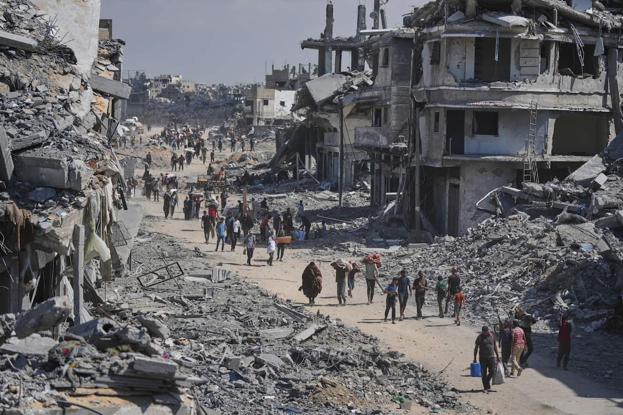 A street is filled with people walking amongst the rubble and heavily damaged buildings of a war-torn urban area.