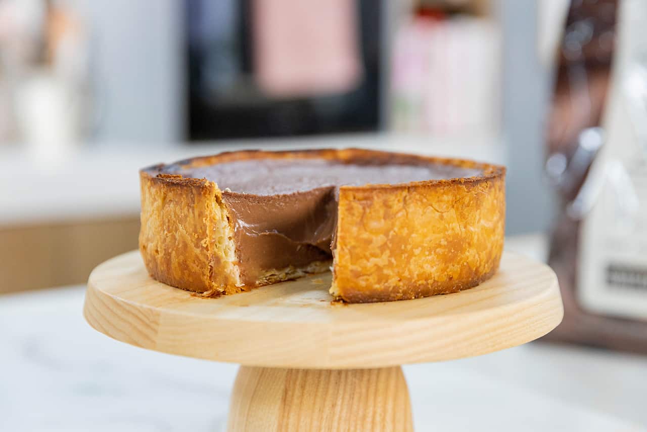 A deep tart with a dark golden-brown pastry case sits on a light-coloured wooden cake stand. A piece has been cut and removed from the flan, showing the baked chocolate custard filling.