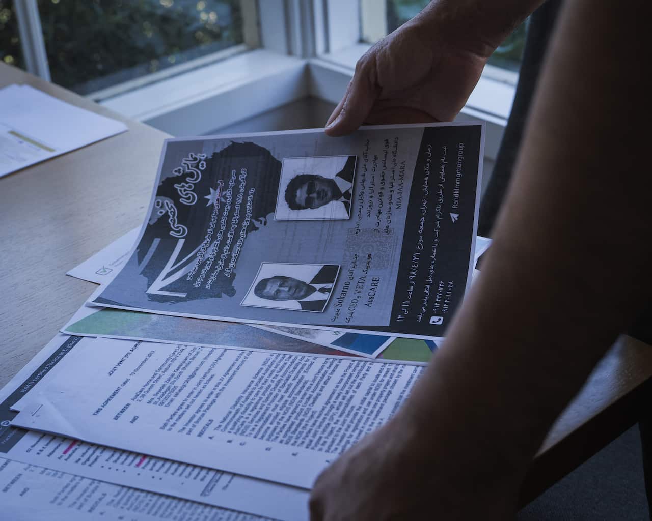 A person's hands are holding up a flyer with two headshots and text in both English and what appears to be Persian. The flyer is part of a stack of documents, including a joint venture agreement, laid out on a table near a window.