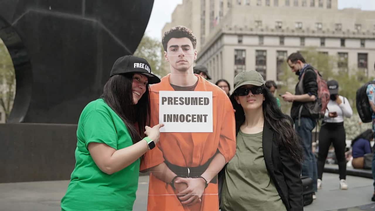 Two women pose for a photo next to a cardboard cutout of Luigi Mangione, who is accused of killing health insurance executive Brian Thompson. Mangione wears an orange jumpsuit, he has his hands tied and on his chest a sign reads 'Presumed Innocent'.