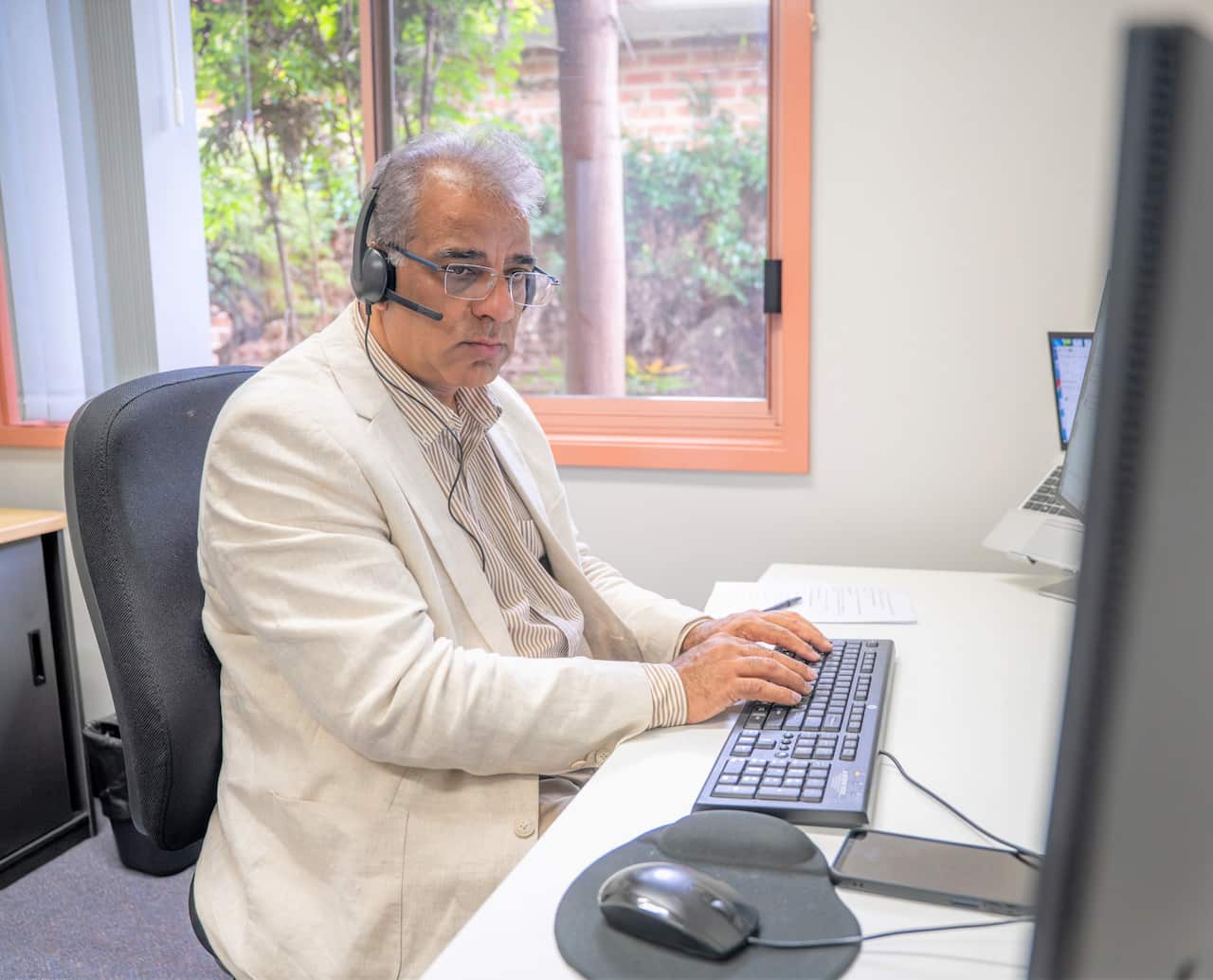 A man in a beige jacket sits at a computer typing on a keyboard.