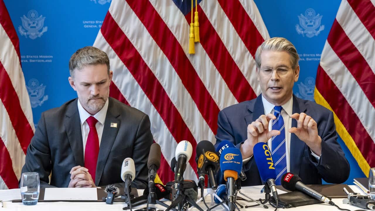 Two men in suits speak into microphones as they sit behind a table with American flags behind them.