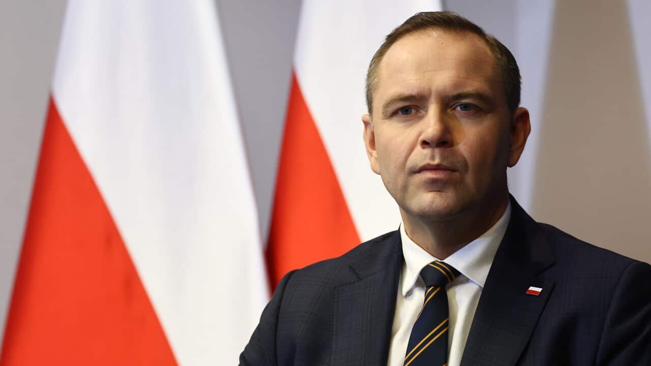 A man in a suit, wearing a Polish flag pin, and standing in front of Polish flags.