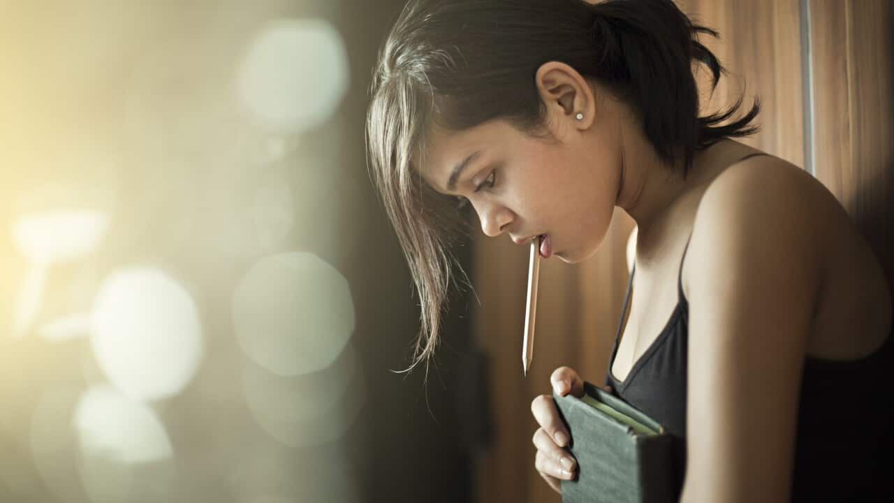 Girl looking down and thinking while holding book and pencil.
