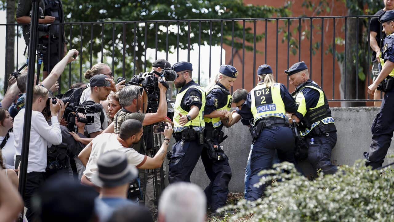 Swedish police officers intervening and detaining a man who set fire to a Quran outside of a mosque while onlookers watch.