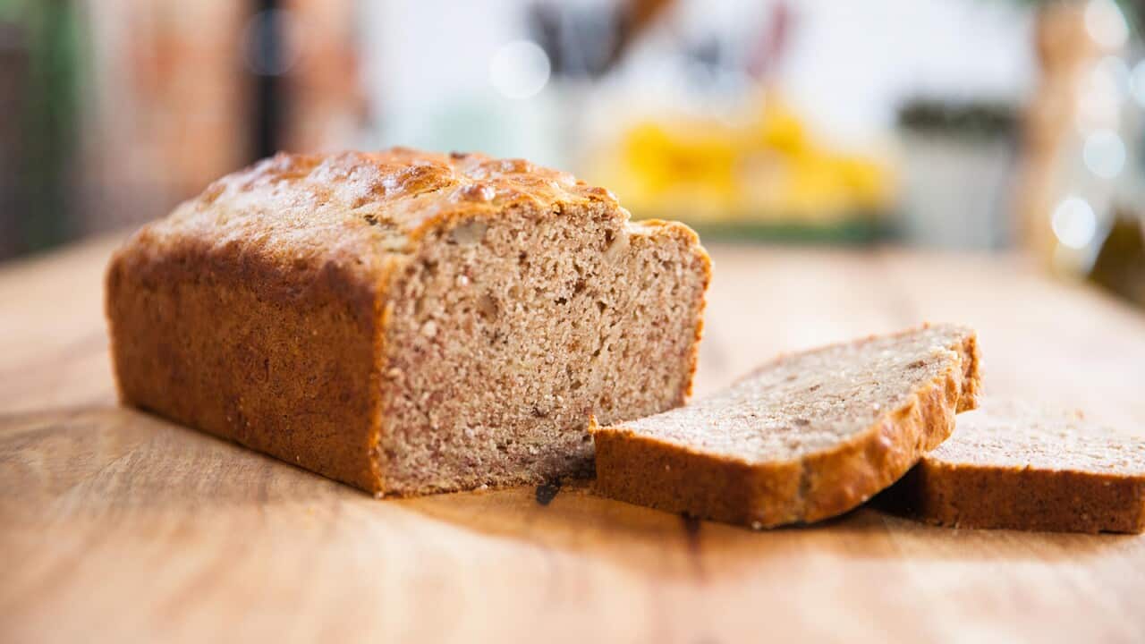 A loaf of banana bread sits on a wooden surface. A few slices have been cut and lie in front.