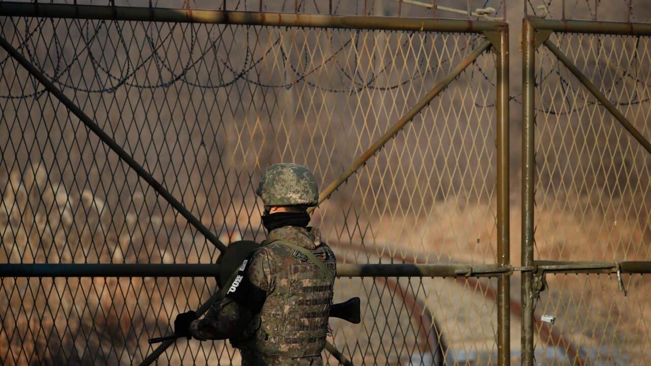 A soldier stands looking out from behind a fence wearing a helmet with a gun in hand