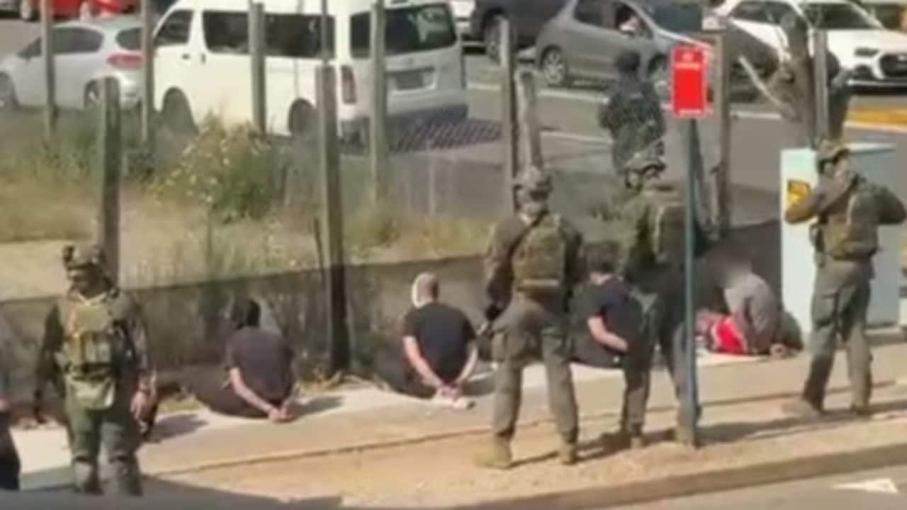 Heavily armed police in camoflauge uniforms stand beside men in who are seated on a footpath with their zip-tied behind their backs