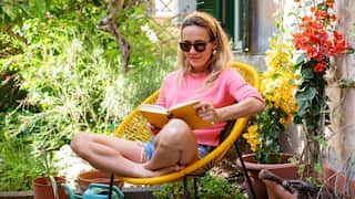 A woman in a pink sweater and sunglasses sits cross-legged in a yellow chair, reading a book in a sunlit garden filled with potted plants and vibrant flowers.