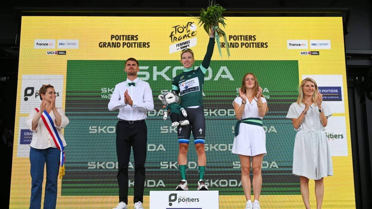 A woman in a cycling kit is standing on a podium holding a bouquet in one hand that she has raised in a sign of victory, and a plushy in the other hand. Four people standing next to her are clapping.