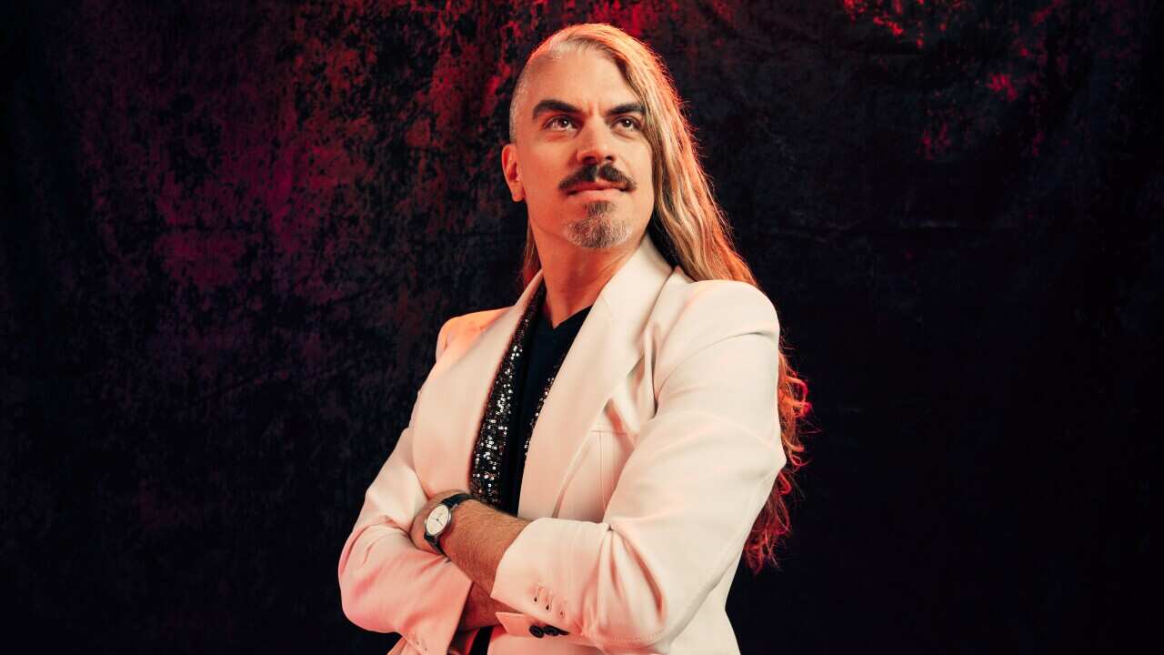 A man in a white blazer with long hair stands with his arms folded