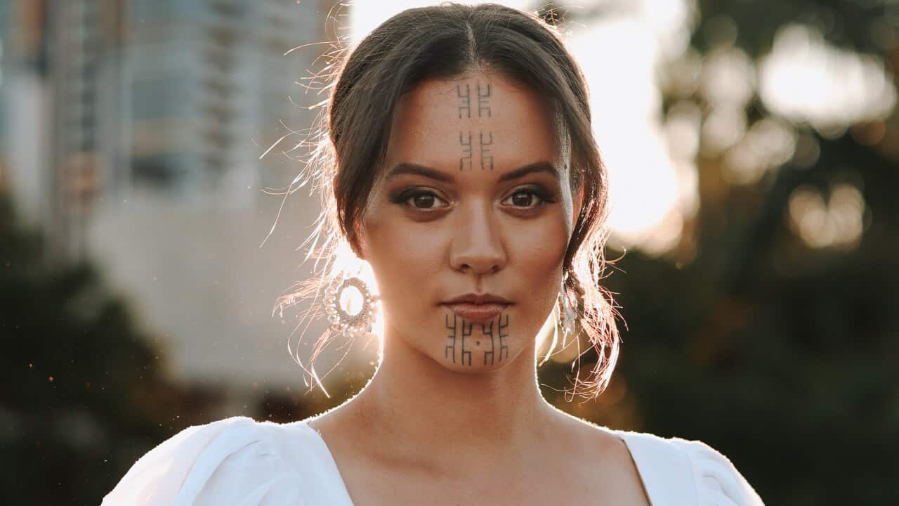 A woman with markings on her face looks at the camera with the sun behind her