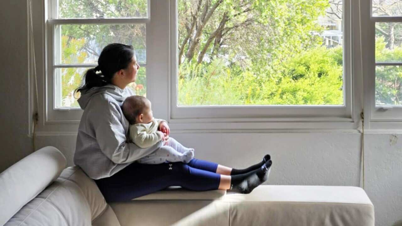An Asian Australian woman in a grey hoodie and blue leggings sits on a sofa looking out the window of her home with her baby propped up in her lap.
