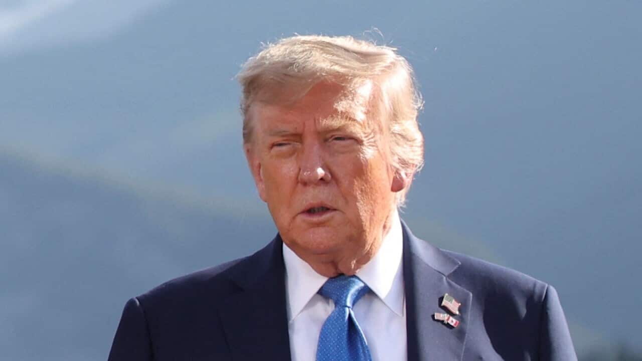 Donald Trump, wearing a suit, white shirt and blue tie. He is wearing two flag pins on his suit, an American flag pin and one showing the American and Canadian flags criss-crossed.