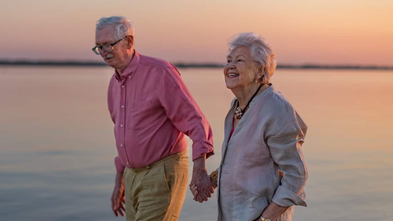 A senior couple walks hand-in-hand along the shoreline of a body of water.