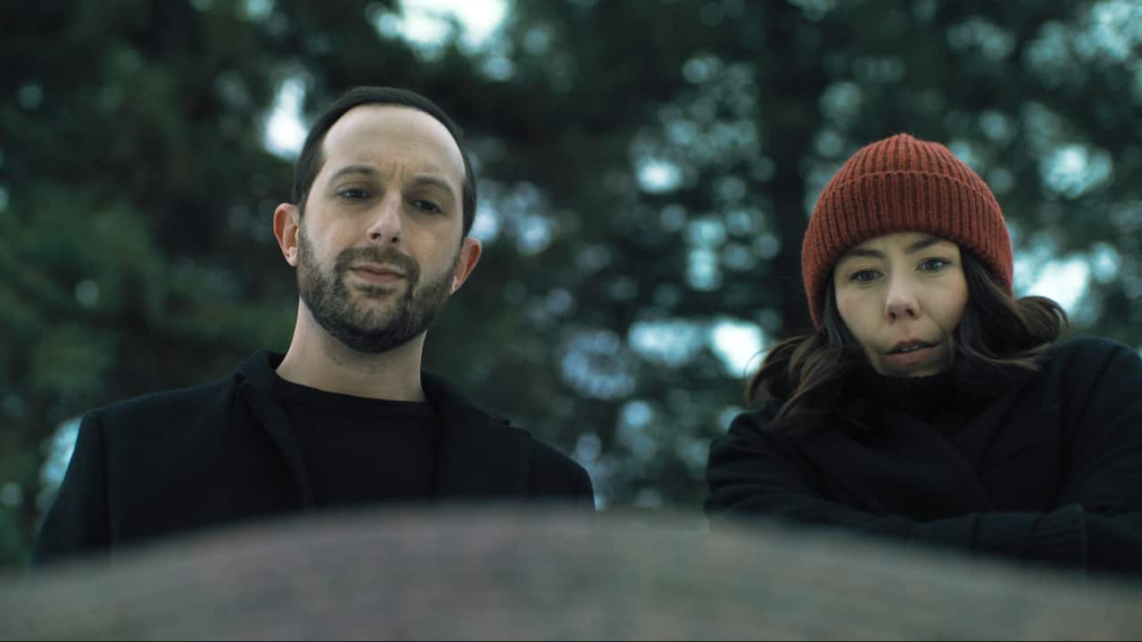 A man and a woman peer down at something out of focus in front of them. The woman is wearing a beanie and the man a coat, suggesting the weather is cold.