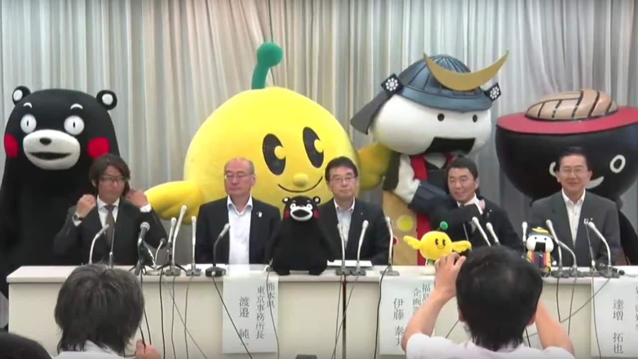 Officials sit behind a table at a press conference, behind them stand four colourful mascots.