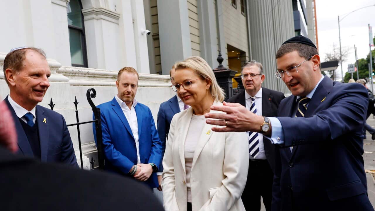 A woman in a white shirt and blazer being introduced to someone by a man outside a synagogue. Four other men are standing nearby.
