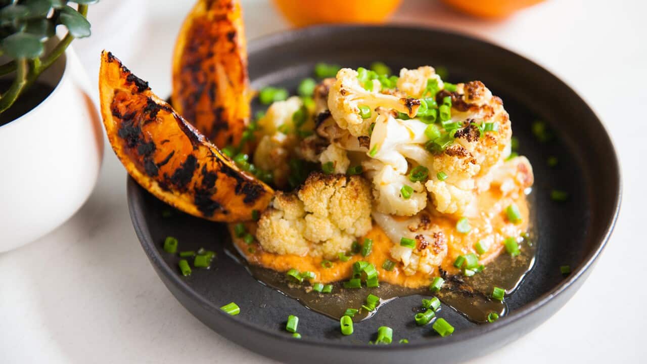 A shallow wide bowl holds pieces of cooked cauliflower sitting on a bed of a pale creamy spread. Two blackened orange wedges sit on one side. The bowl is sprinkled with chives.