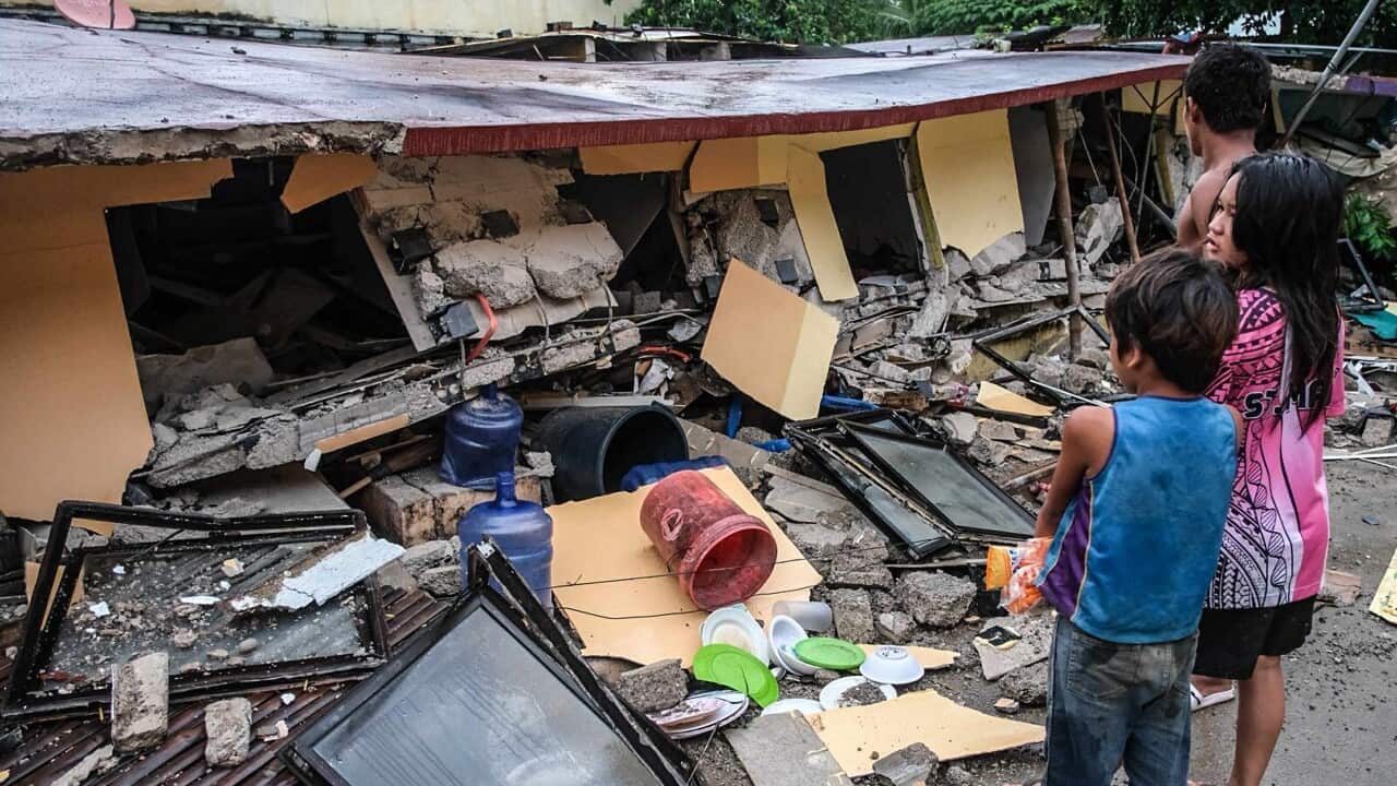 Three people, including two children, look at a collapsed house after an earthquake.