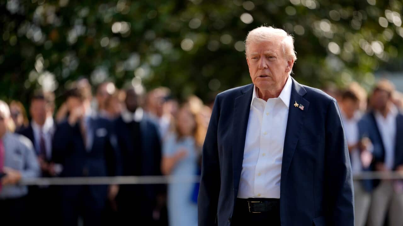 A close-up photograph features Donald Trump walking outdoors, dressed in a navy suit jacket, dark trousers, and an unbuttoned white shirt, holding what appears to be a white hat in his right hand.
