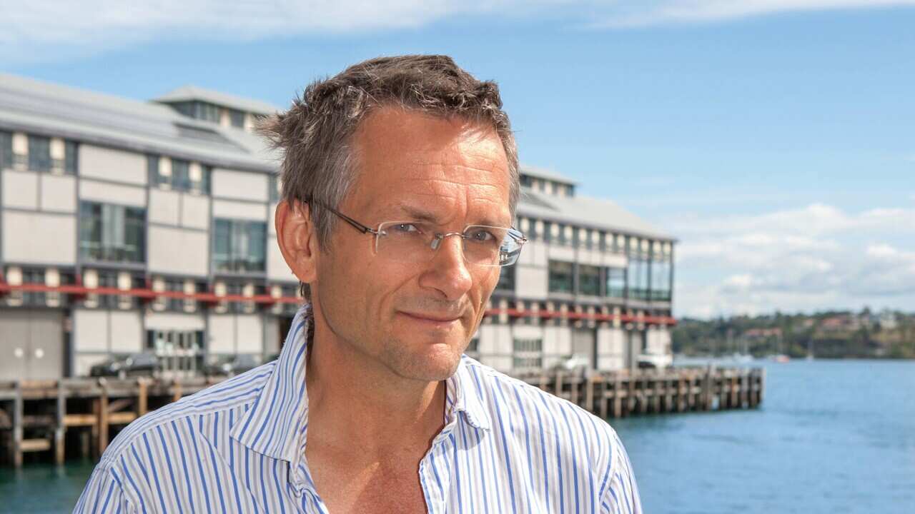 A man wearing a striped collared shirt stands with his hands in his pockets against the backdrop of water.