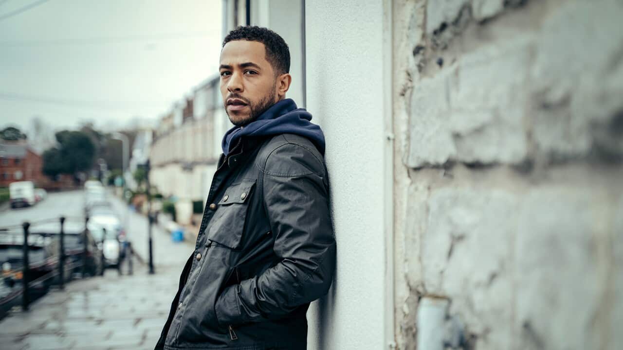 A man in blue hoodie under a dark jacket leans against a pale stone wall, looking sideways at the camera. A street stretches away behind him.
