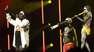 An Aboriginal Australian singer in a long-sleeved white shirt points his finger upwards on a dark stage with red and yellow lights, while three men in body paint and red and yellow loincloths perform beside him, one playing a didgeridoo.