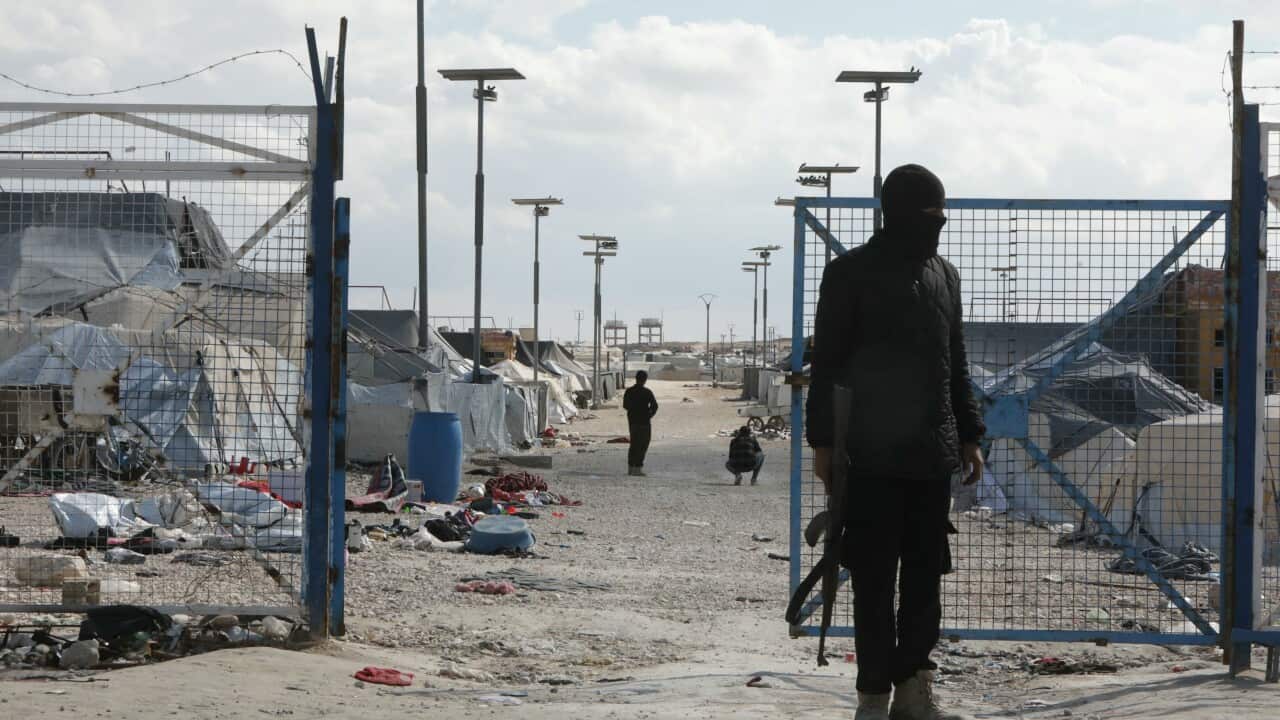 An armed, masked guard stands outside the gates of the Al-Hol detention camp in north eastern Syria. The gates are open and littered belongings surround the tents inside. In the background, two more guards survey the scene inside the camp.