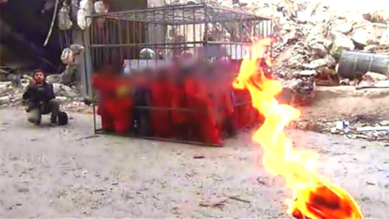 A man crouches by a metal cage containing children dressed in orange. A flamed torch is in the foreground. 