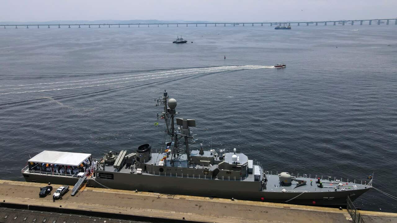 An aerial view shows a grey naval corvette docked at a pier with a reception tent on its aft deck, while a long bridge stretches across the water in the background.