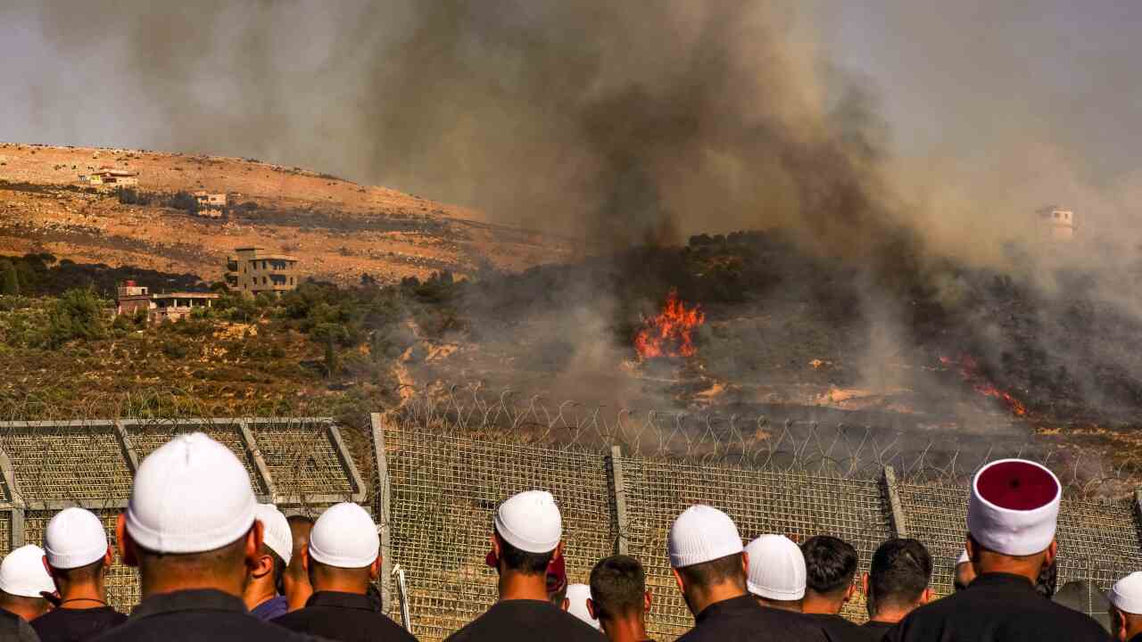 A group of people stand with their backs to the camera looking at a fire in the distance