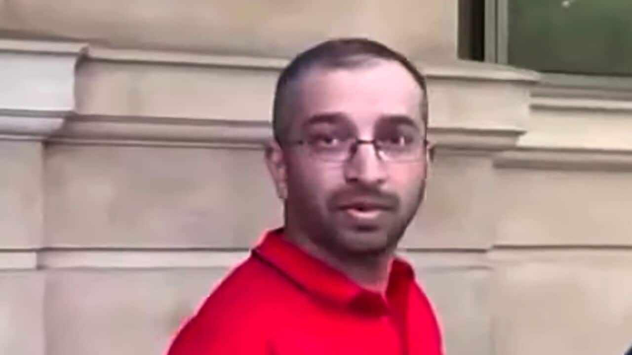 A man in a red t-shirt, holding papers in one hand, looks on.