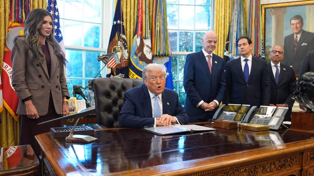 Donald Trump sitting in his chair in the Oval Office, flanked by a woman in a brown blazer, and three men in navy blazers.