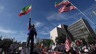 A man holding an Iranian flag high above his head stands above a crowd holding a mixture of Iranian and American flags