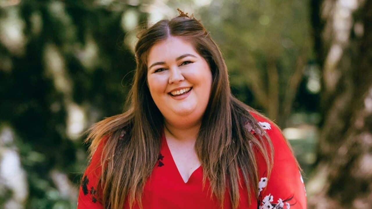 a young woman with long brown hair and in a red dress with white flowers smiles at the camera in front of a blurred background of a green park