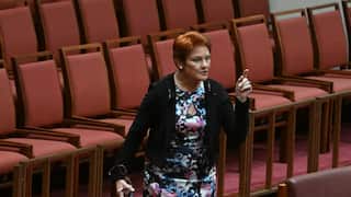 Pauline Hanson, wearing a floral dress, gesticulating in a room with red seats and pink carpet.