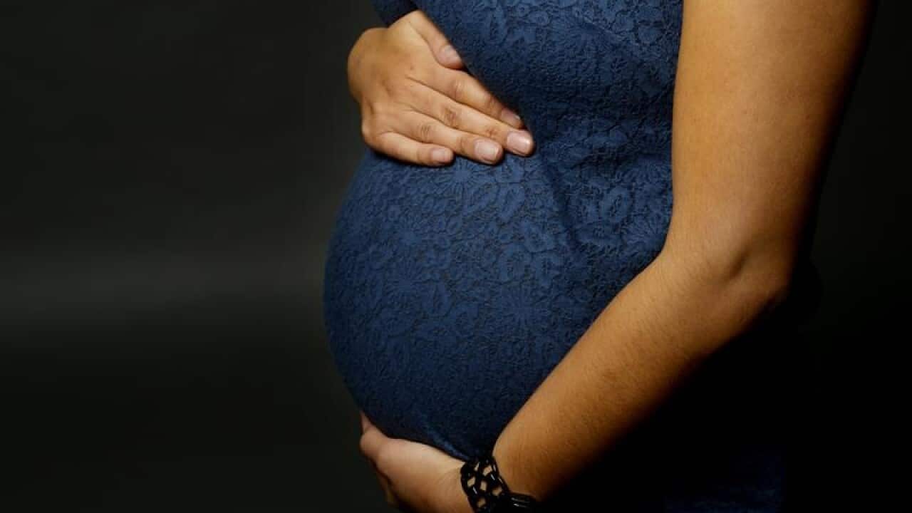 A pregnant woman's torso. She is wearing a blue dress and has one hand at the top of her belly and one below.