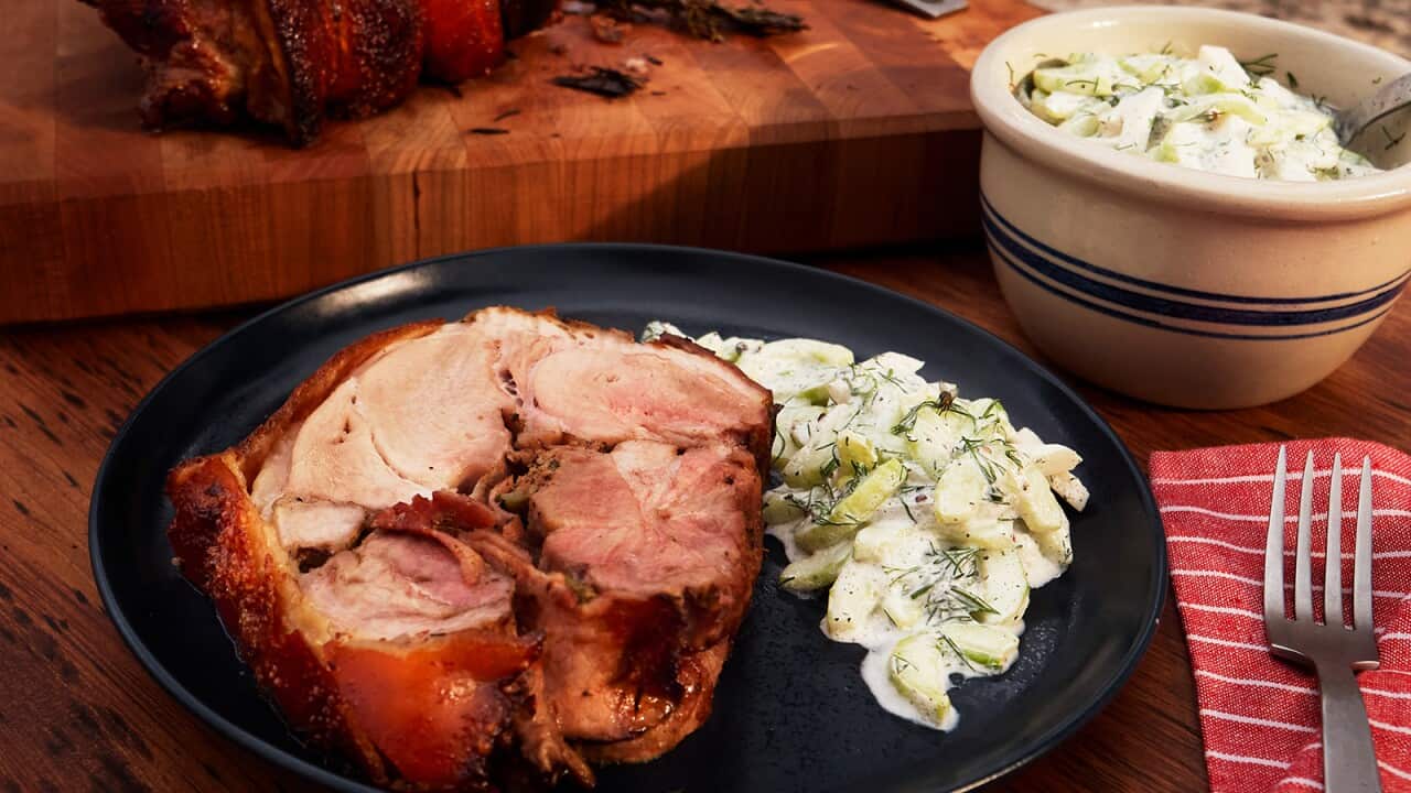 A thick slice of meat and some creamy salad sits on a round plate on a wooden surface, with a striped red napkin and a silver fork alongside.