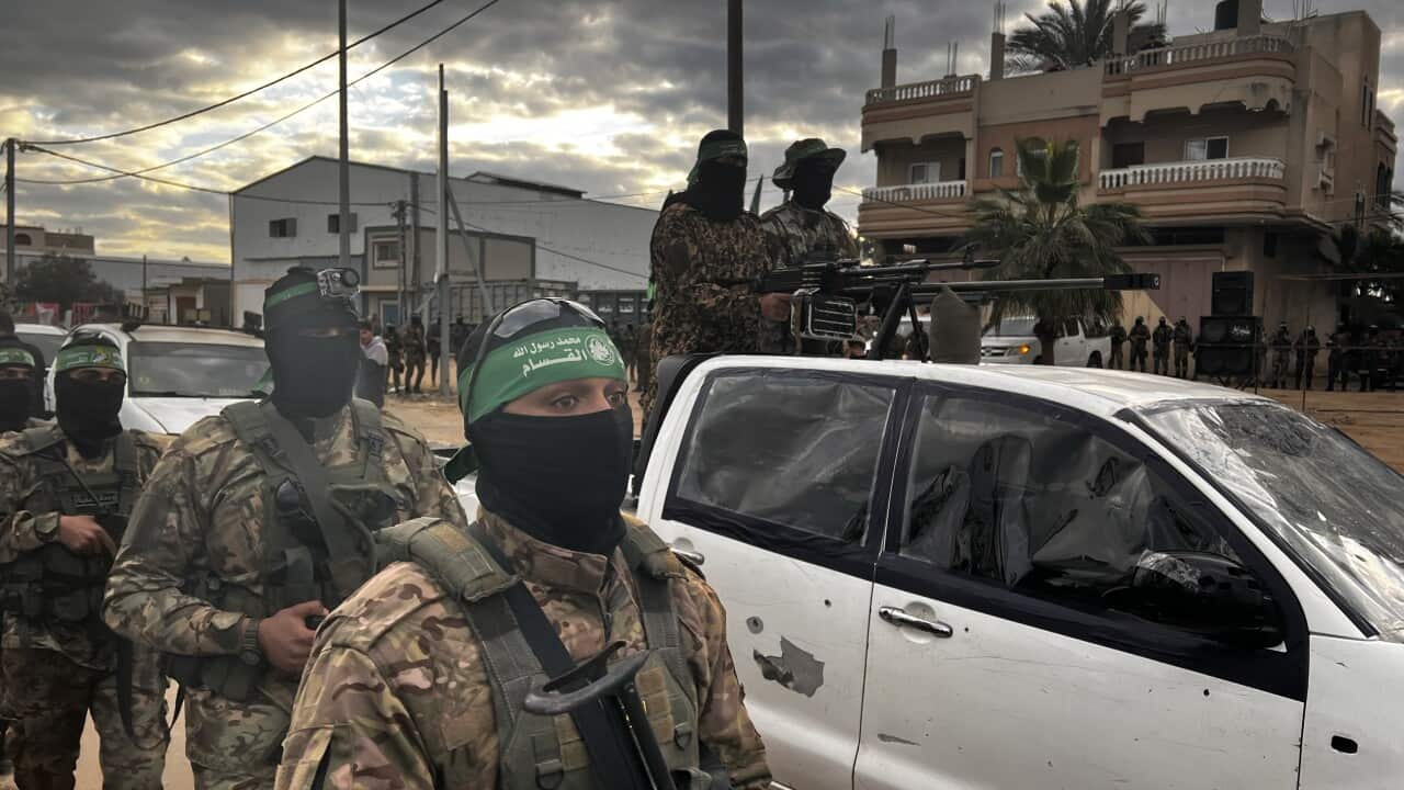 Several men wearing military uniforms and black balaclavas walk next to a white utility vehicle, in the tray of which stand two similarly clad men holding a mounted automatic rifle.