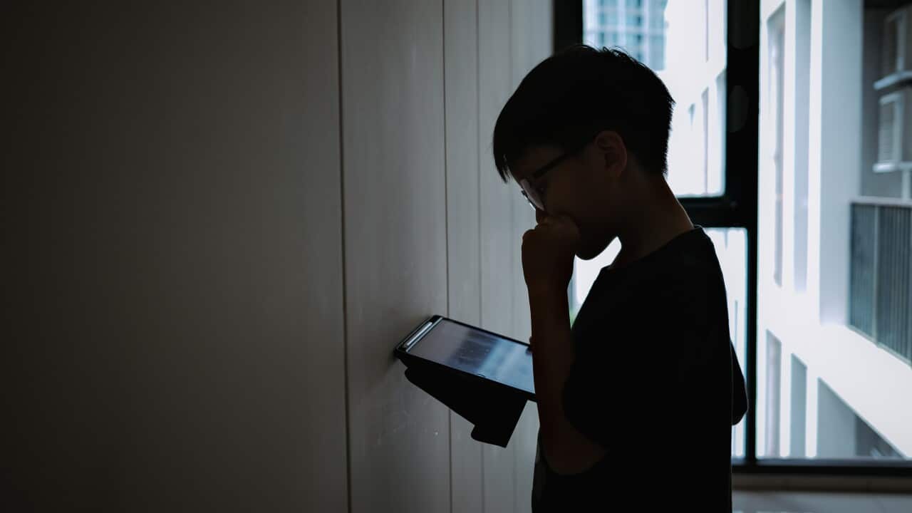 Silhouette photo of a young boy holding and looking at computer tablet against the wall inside an apartment alone.