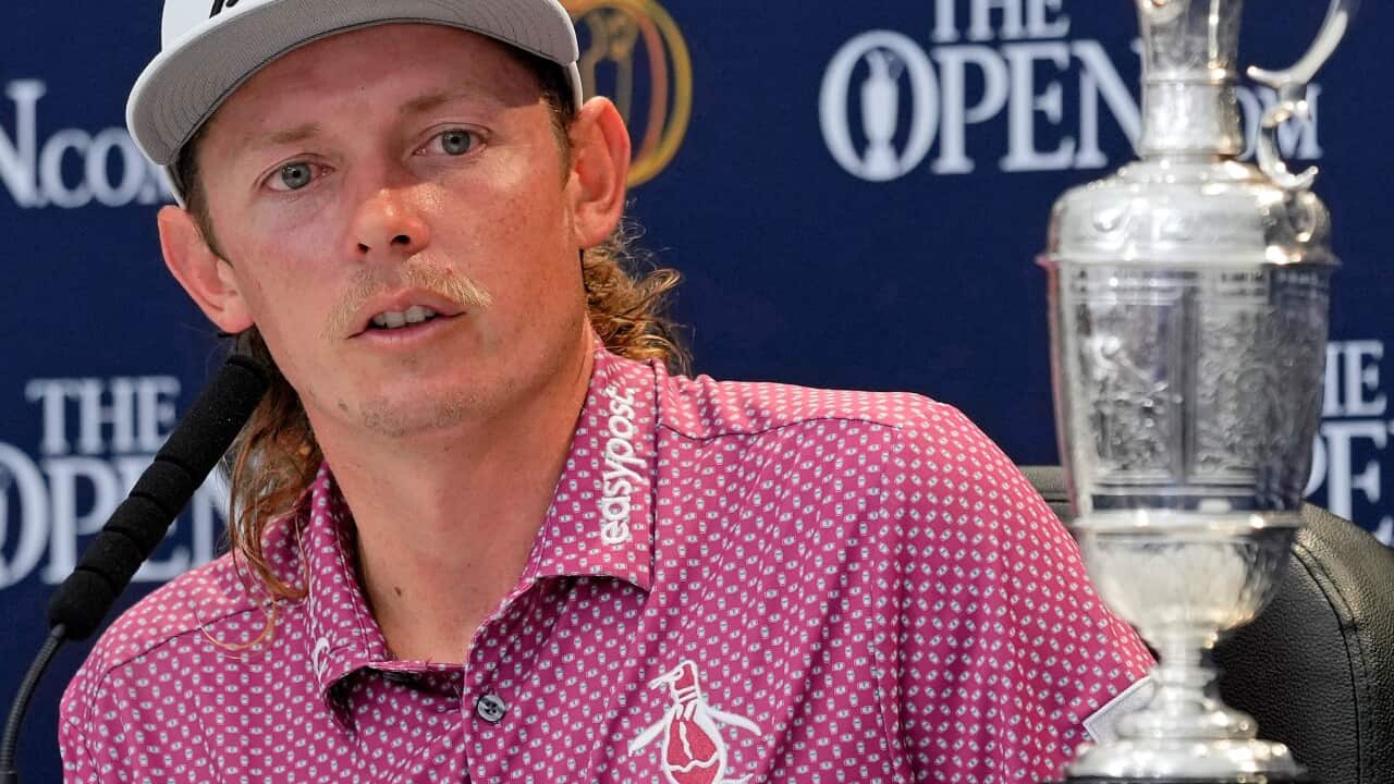 A man sitting while speaking into a microphone. A silver trophy sits next to him on a table.