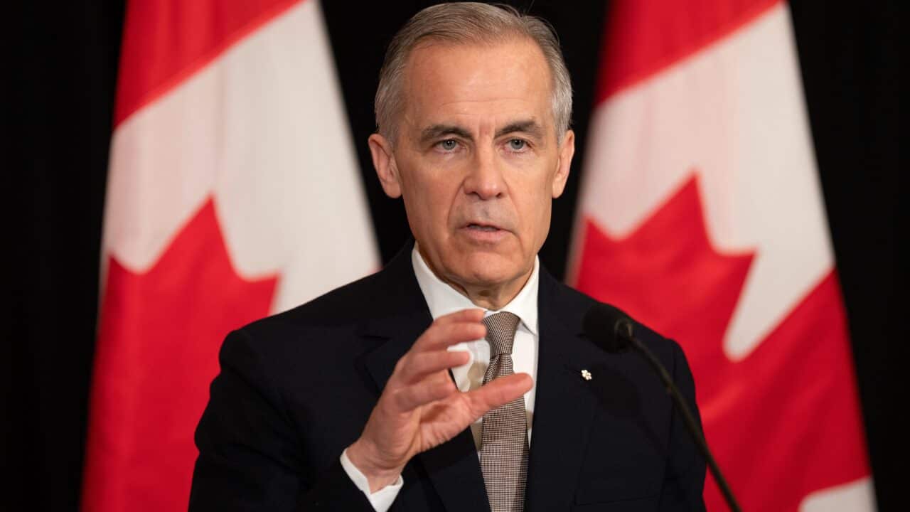 A grey-haired man gestures with his hand. With two canadian flags blurry in the background.
