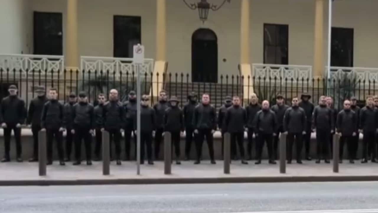 A group of black-clad men stand with their arms by their side in front of the NSW Parliament.