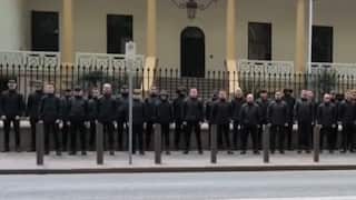 A group of black-clad men stand with their arms by their side in front of the NSW Parliament.