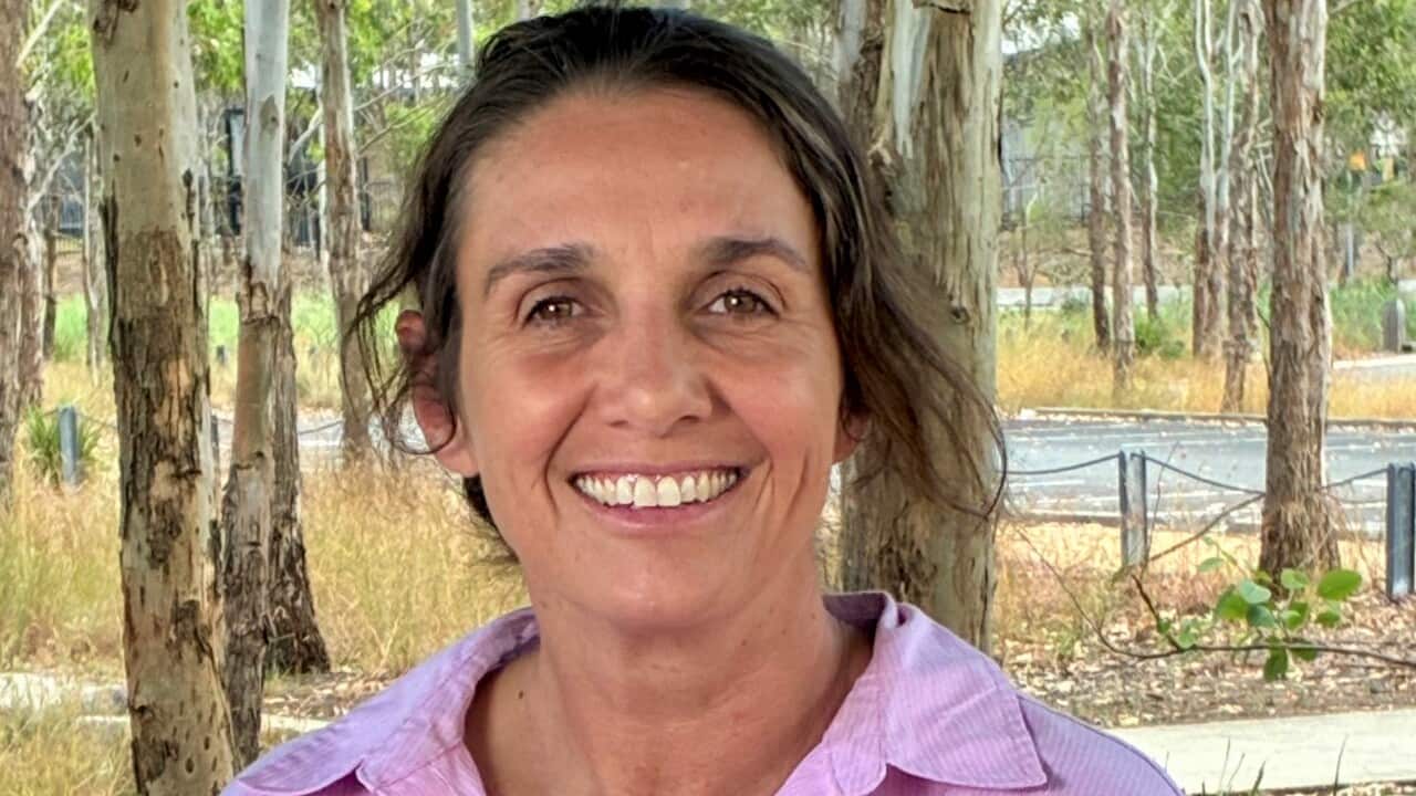 A woman in a pink shirt sits in a bushland hut, with arms folded on a table.