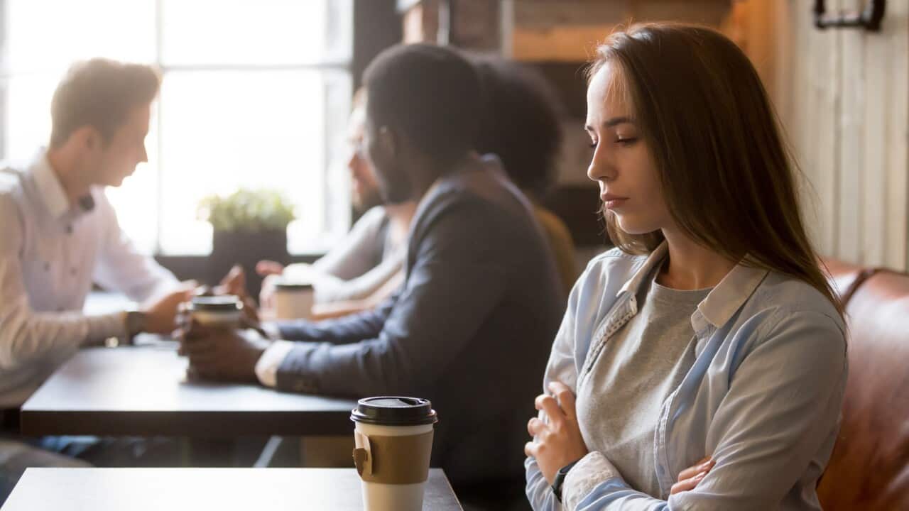 A young girl with long hair sits alone in a coffee shop with a crowded table of people behind her. She looks downcast.