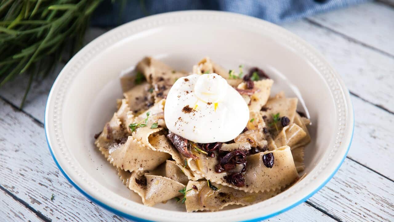 A white bowl with a blue rim sits on a white-washed wooden surface. The bowl holds ribbons of pasta, topped with a round of mozzarella.