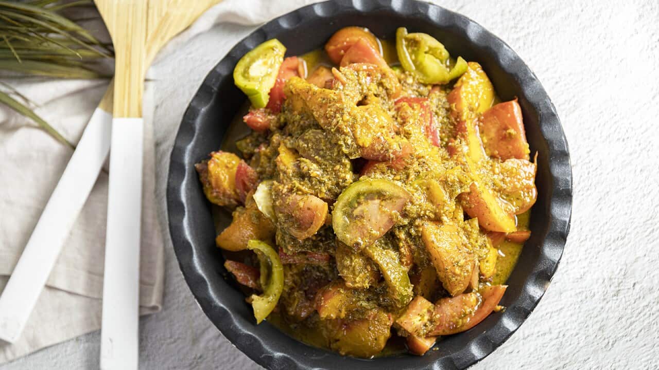 A tomato and peach salad, with a thick sauce, sits in a dark bowl on a white surface. Salad servers sit alongside, on a napkin.
