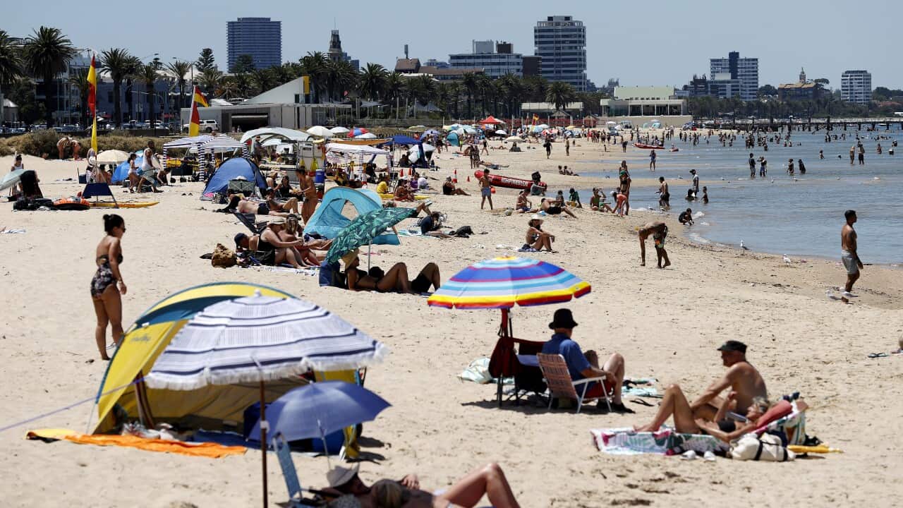 A beach scene featuring multiple people enjoying the sun, with some standing, others lying on towels, some sitting in beach chairs, and a few wading in the water.